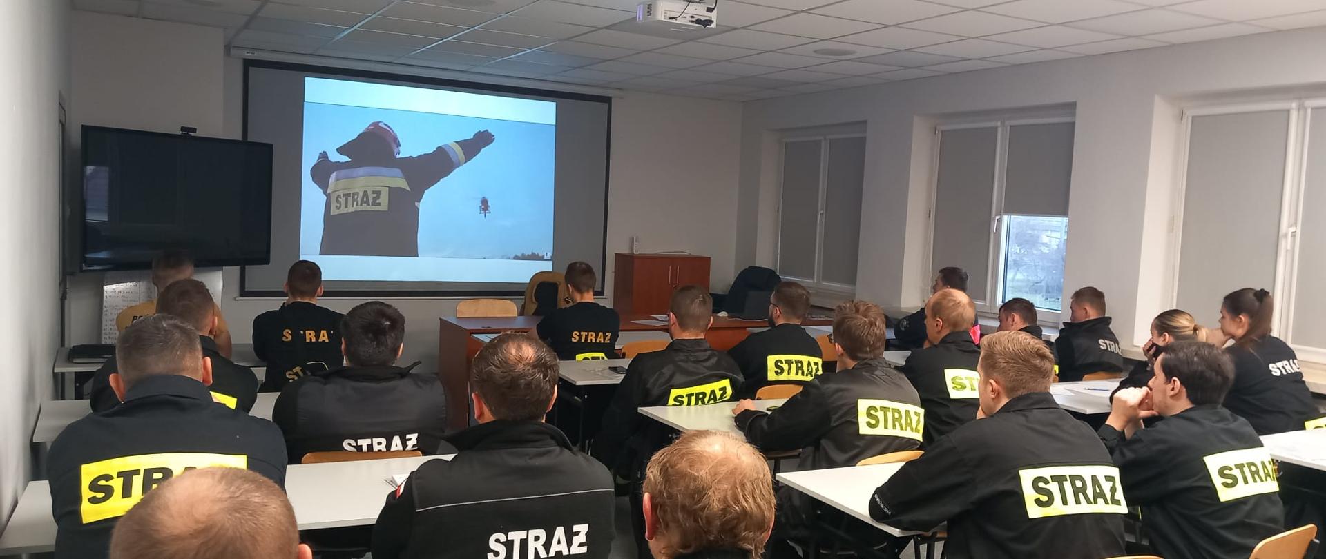 Na sali wykładowej Ośrodka Szkolenia Komendy Wojewódzkiej Państwowej Straży Pożarnej w Opolu, kursanci, strażacy OSP i PSP siedzą i oglądają film szkoleniowy. Strażacy w umundurowaniu koszarowym oraz służbowym.