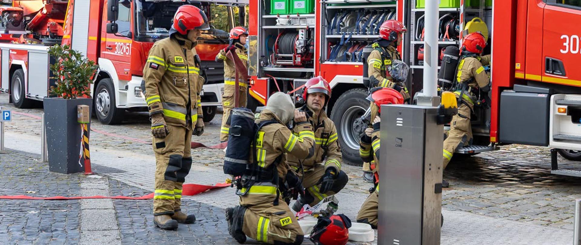 Strażacy w ubraniach specjalnych i czerwonych hełmach przygotowują sprzęt ratowniczy na placu przed wozami bojowymi. Z tyłu widoczne są otwarte skrytki sprzętowe czerwonego samochodu ratowniczego oraz drabina mechaniczna. 