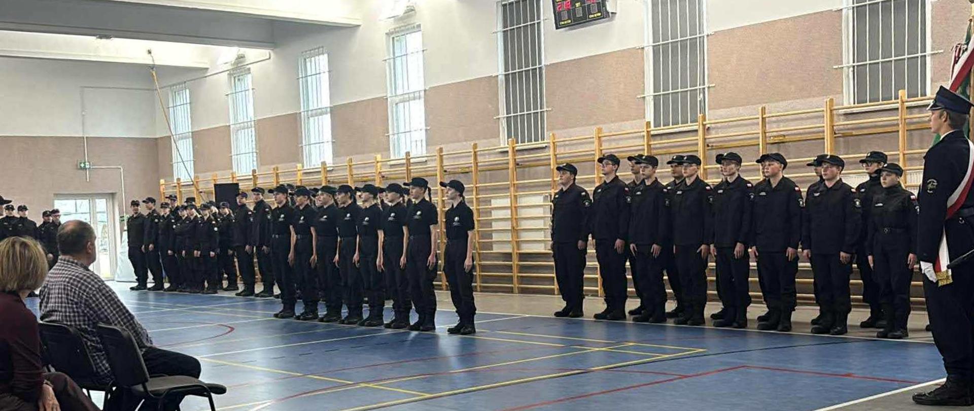 Uczniowie i uczennice w czarnych mundurach ustawieni oddziałami w dwuszeregu w sali gimnastycznej. Obok stoi poczet sztandarowy. Naprzeciwko siedzi publiczność.