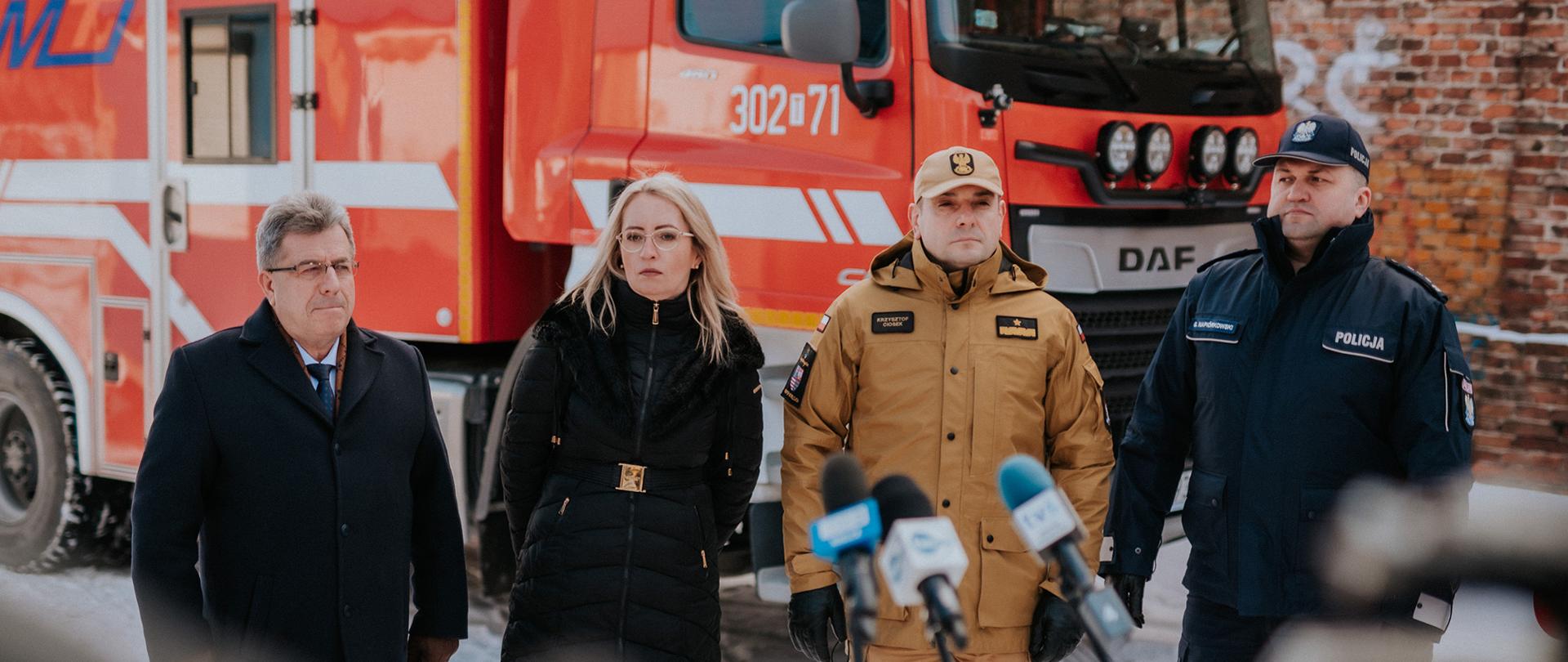 Na zdjęciu widzimy uczestników konferencji prasowej od lewej w garniturze i płaszczu mężczyzna, potem kobieta w kurtce zimowej ciemnej, potem strażak w mundurze piaskowym i policjant w mundurze granatowym. Stoją na dworze, dookoła śnieg a w tle samochód strażacki. 