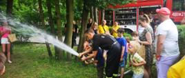 Na zdjęciu widać strażaka i chłopca podających prąd wody z prądownicy wodnej typu turbo. W pobliżu znajdują się inne dzieci i ich opiekunowie. W tle widać samochód strażacki i strażaków.