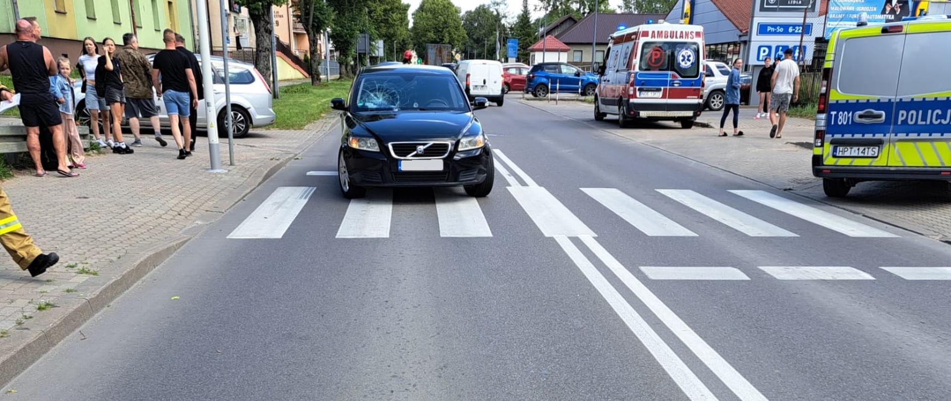 Czarny samochód osobowy, kombi, marki volvo, który stoi na jezdni, na pasach, na których doszło do potrącenia. Po prawej stronie na chodniku stoi radiowóz Policji. W tle widoczna jest karetka ZRM oraz pojazdy osobowe i gapie.