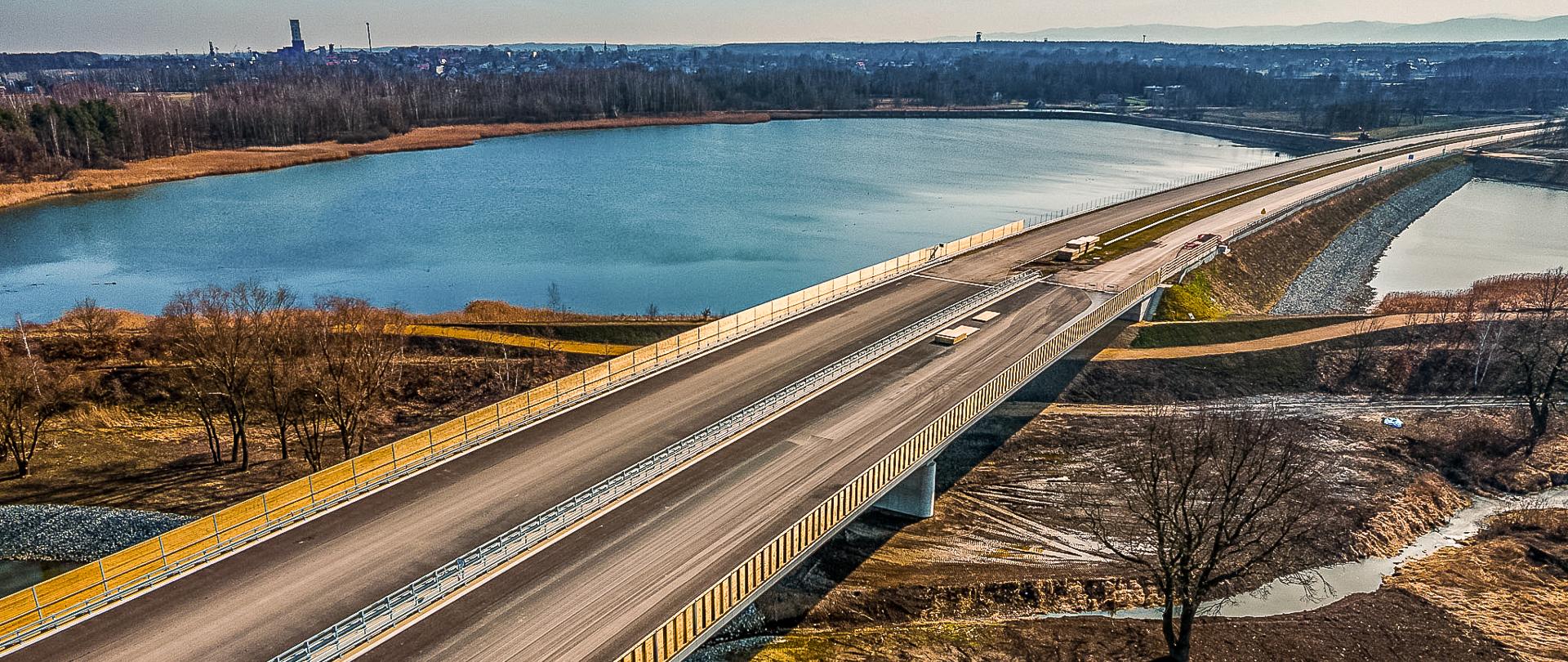 Fotografia ukazuje imponujący most rozpościerający się nad zbiornikiem wodnym. Most wyróżnia się nowoczesnym designem, z szeroką jezdnią otoczoną barierkami ochronnymi. Kamieniste brzegi pod nim przechodzą w spokojną taflę wody. W tle malowniczo rozciąga się duży zbiornik wodny otoczony zielenią, a na horyzoncie można dostrzec miejskie budynki oraz wieżę. Sceneria sugeruje harmonijne połączenie natury z architekturą.