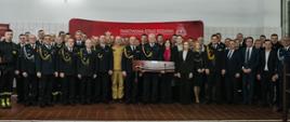 Pamiątkowe zdjęcie grupowe funkcjonariuszy Państwowej Straży Pożarnej oraz zaproszonych gości wykonane w hali jednostki. Kilkadziesiąt osób w umundurowaniu galowym i strojach oficjalnych stoi w kilku rzędach na tle czerwonego baneru Komendy Powiatowej PSP we Włoszczowie; w centrum widoczny oficer trzymający symboliczny upominek. Atmosfera uroczystego zakończenia wydarzenia służbowego.