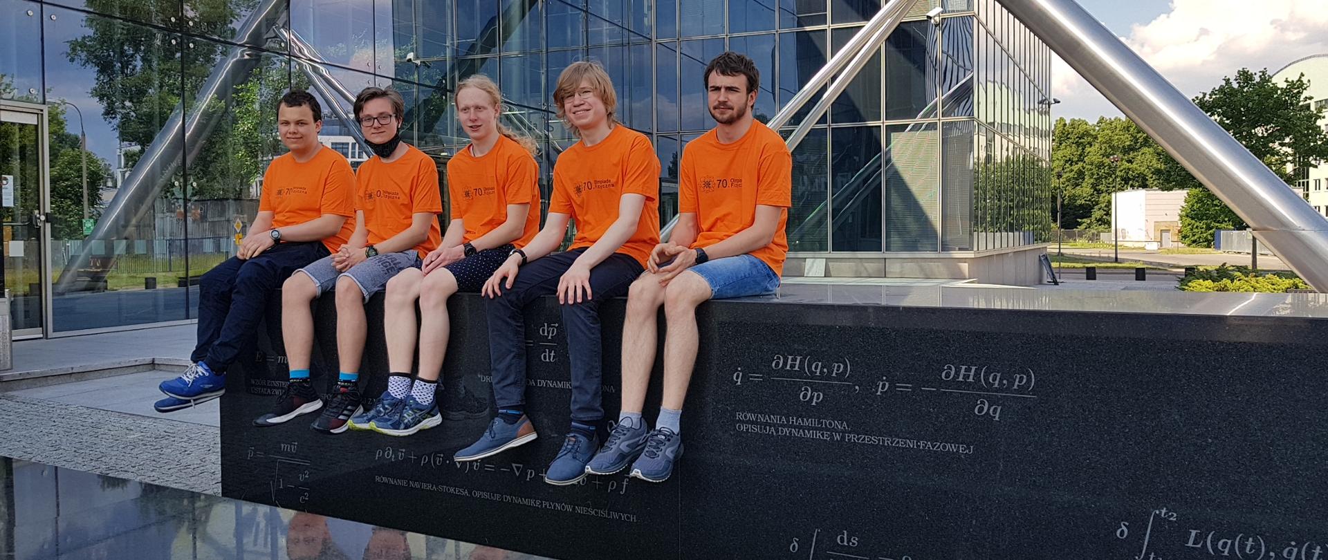 Pięcioro olimpijczyków siedzących pod dużym przeszklonym budynkiem.