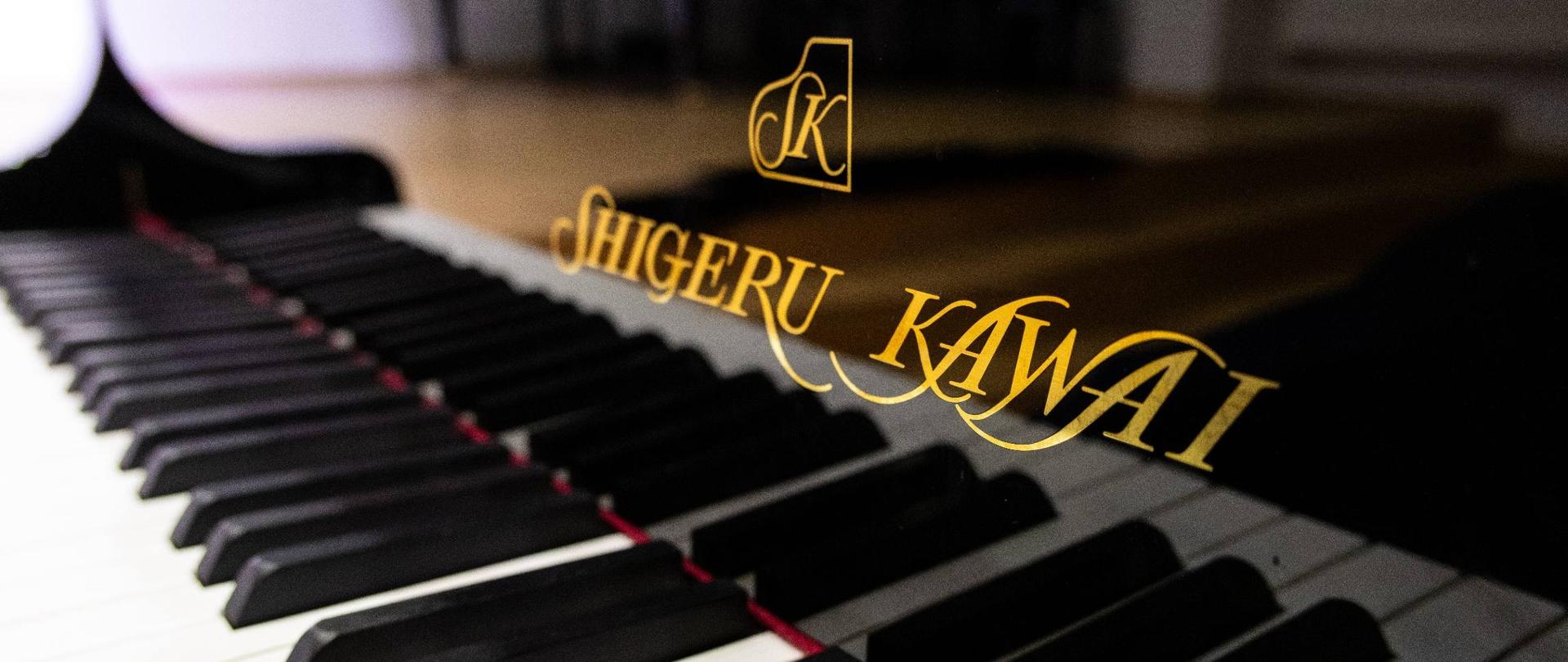 Zdjęcie przedstawia klawiaturę nowego fortepianu Shigeru Kawai SK-EX ze złotym napisem i logo firmy na froncie.