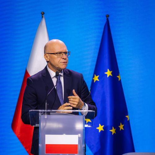 Ambasador Rafał Wiśniewski stojący przy podium na tle flag. 