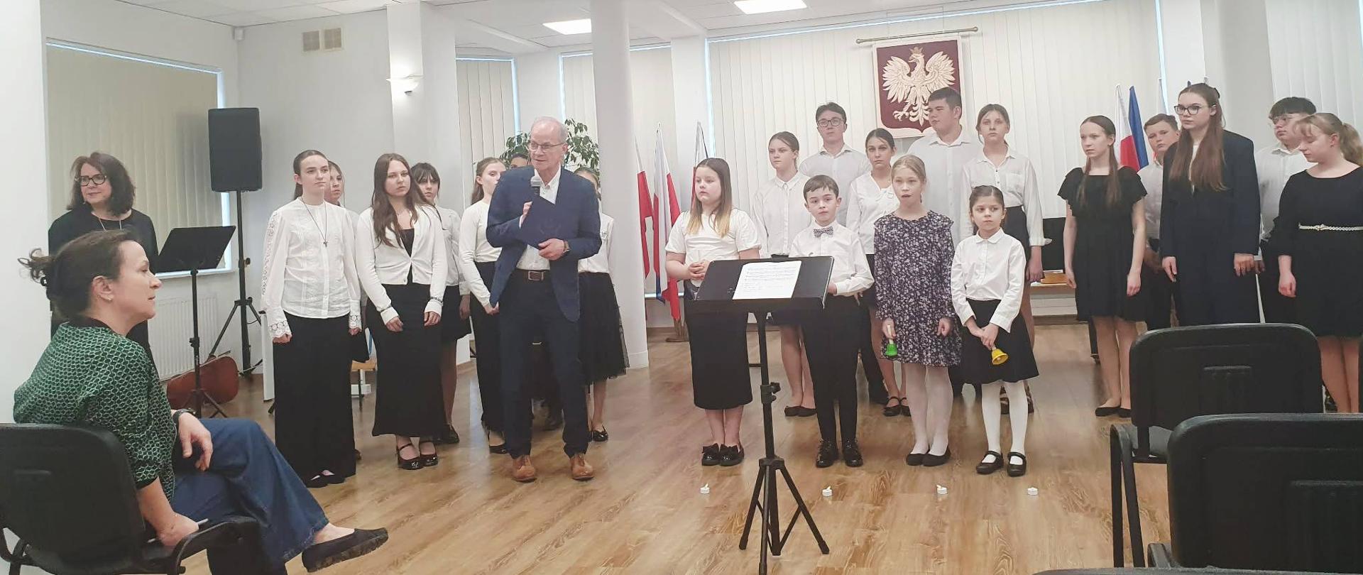 W sali stoi grupa dziewczyn i chłopców. Dzieci ubrane są odświętnie w białe koszule oraz czarne spodnie lub spódnice. Przed nimi stoi mężczyzna ubrany w granatowy garnitur i przemawia przez mikrofon. Zgromadzonych obserwuje kobieta siedząca przed nimi na krześle.