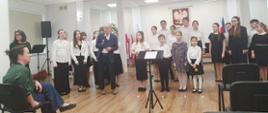 W sali stoi grupa dziewczyn i chłopców. Dzieci ubrane są odświętnie w białe koszule oraz czarne spodnie lub spódnice. Przed nimi stoi mężczyzna ubrany w granatowy garnitur i przemawia przez mikrofon. Zgromadzonych obserwuje kobieta siedząca przed nimi na krześle.