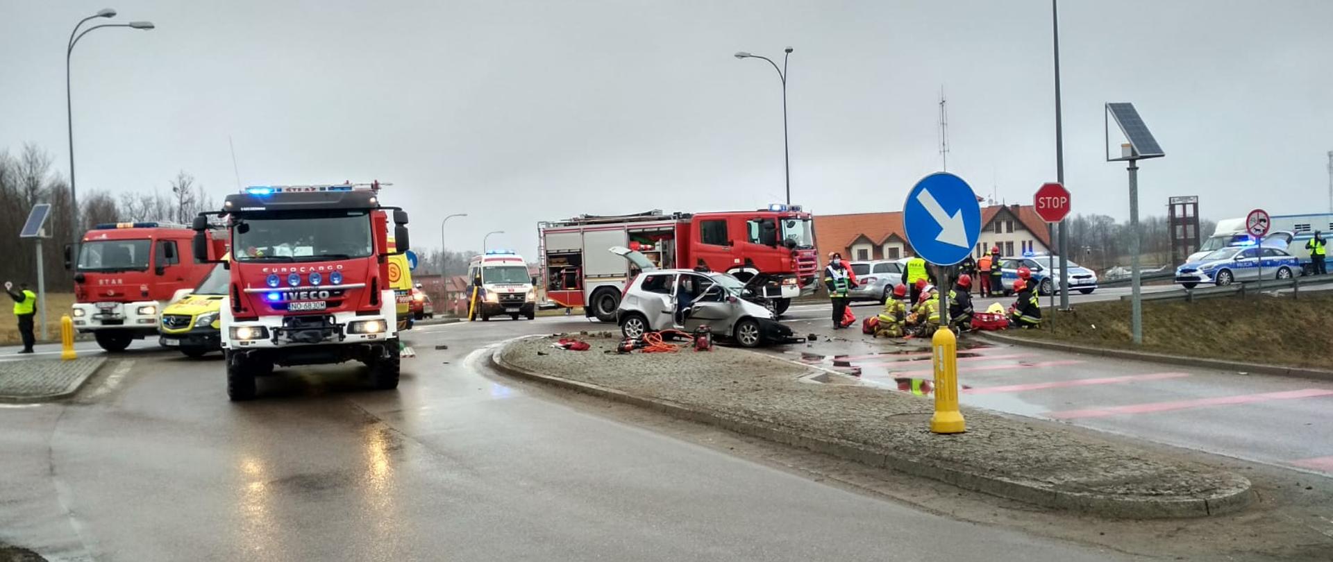Zdjęcie przedstawia miejsce działań po wypadku dwóch samochodów osobowych. Od lewej strony: jednego funkcjonariusza Policji kierującego ruchem drogowym, dwa pojazdy pożarnicze i jedną karetkę pogotowia,. Następnie kolejną karetkę pogotowia oraz w centrum wrak jednego z samochodów osobowych, za którym usytuowany jest kolejny pojazd pożarniczy. Prawa część zdjęcia obrazuje udzielanie pomocy medycznej osobom poszkodowanych przez strażaków (w tle drugi z aut osobowych). Następnie znajdują się dwa patrole Policji i jeden funkcjonariusz