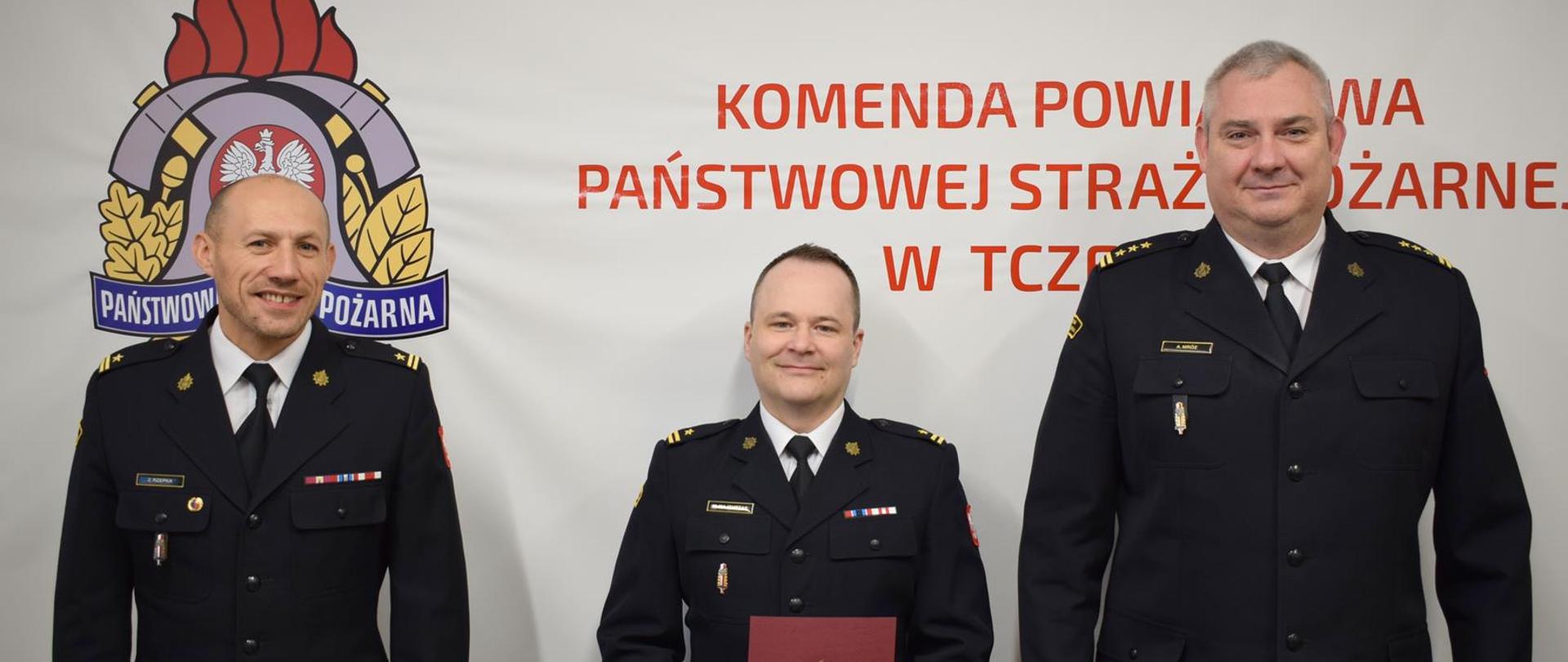 Wspólne zdjęcie Komendanta, Zastępcy oraz mianowanego dowódcy. W tle logo PSP i napis KOMENDA POWIATOWA PAŃŚTWOWEJ STRAŻY POŻARNEJ W TCZEWIE.