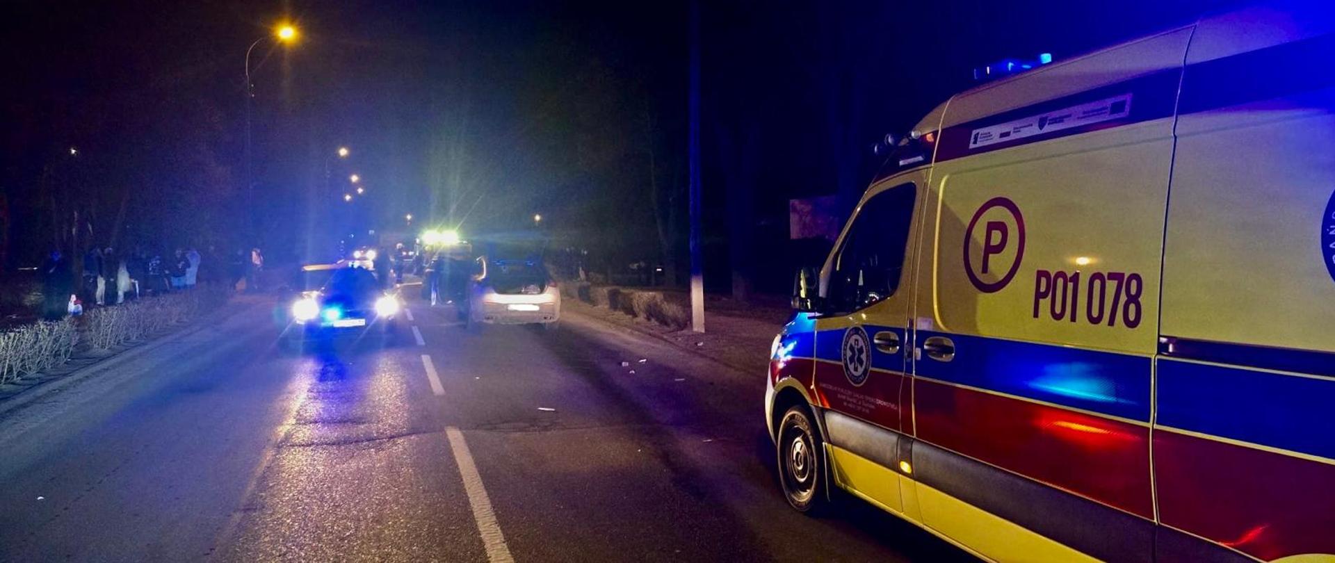 Nocna scena na drodze – po prawej stronie stoi karetka pogotowia z włączonymi niebieskimi światłami, a na środku jezdni widoczny jest radiowóz policyjny oraz zatrzymany samochód osobowy. W tle widać kolejne pojazdy z sygnałami świetlnymi i kilka osób stojących przy poboczu. Ulica jest oświetlona latarniami, a wokół panuje ciemność.