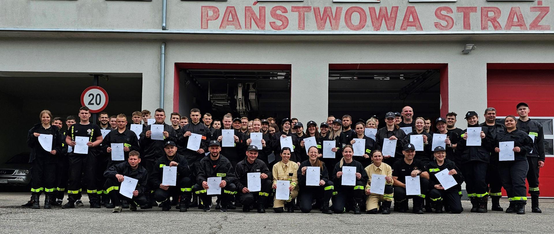 Zbiórka do pamiątkowego zdjęcia strażaków ubranych w piaskowe i czarne mundury na utwardzonym placu. W tle budynek z czerwonymi bramami garażowymi.