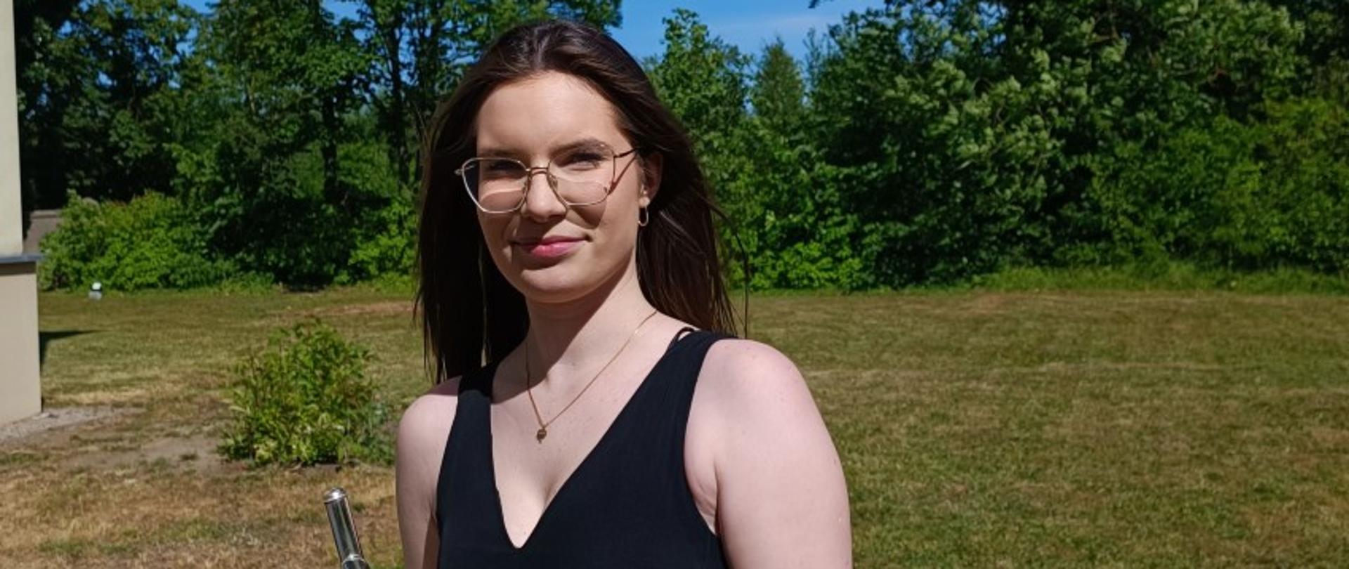 Zdjęcie kolorowe. Młoda flecistka w stroju galowym stoi z instrumentem na tle drzew.