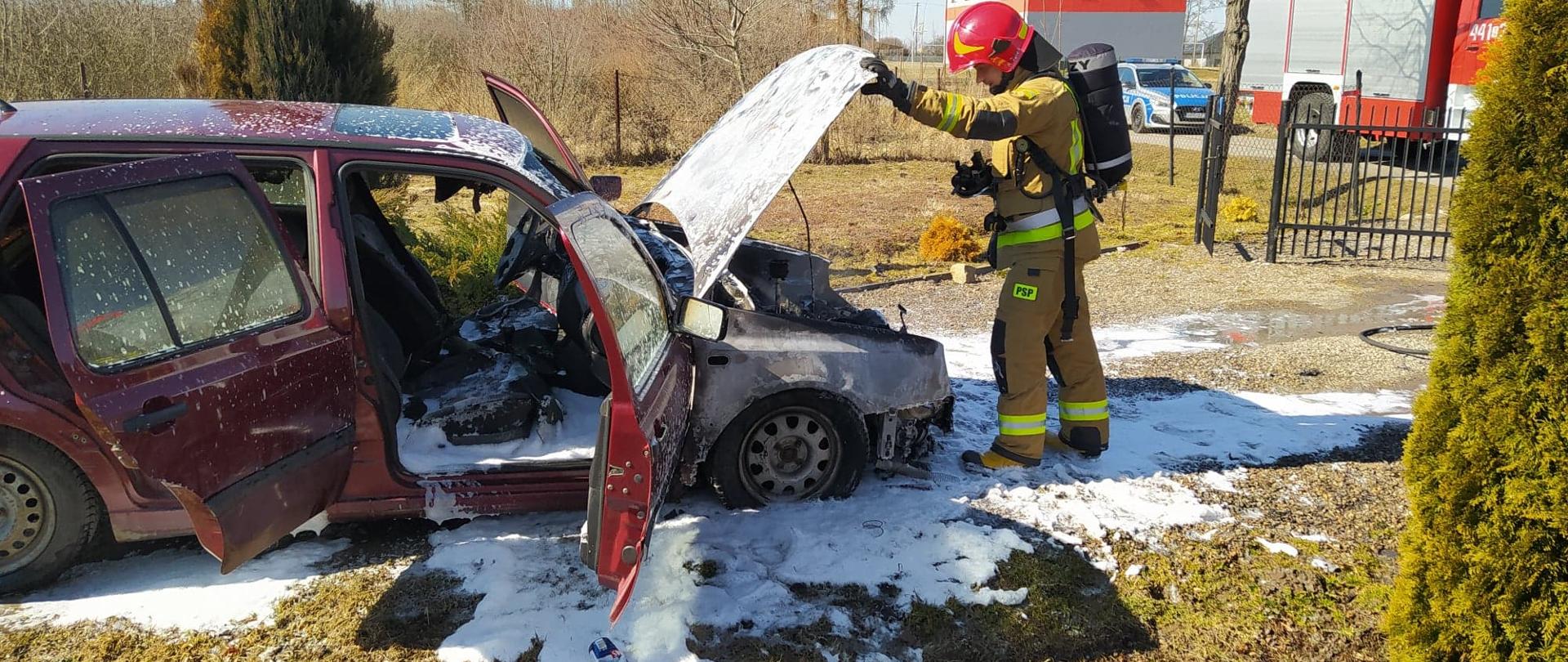 Zdjęcie przedstawia czerwony samochód z otwartymi drzwiami i przednią maską. pod samochodem znajduje się piana gaśnicza przed samochodem stoi strażak w tle pojazd pożarniczy oraz policyjny radiowóz