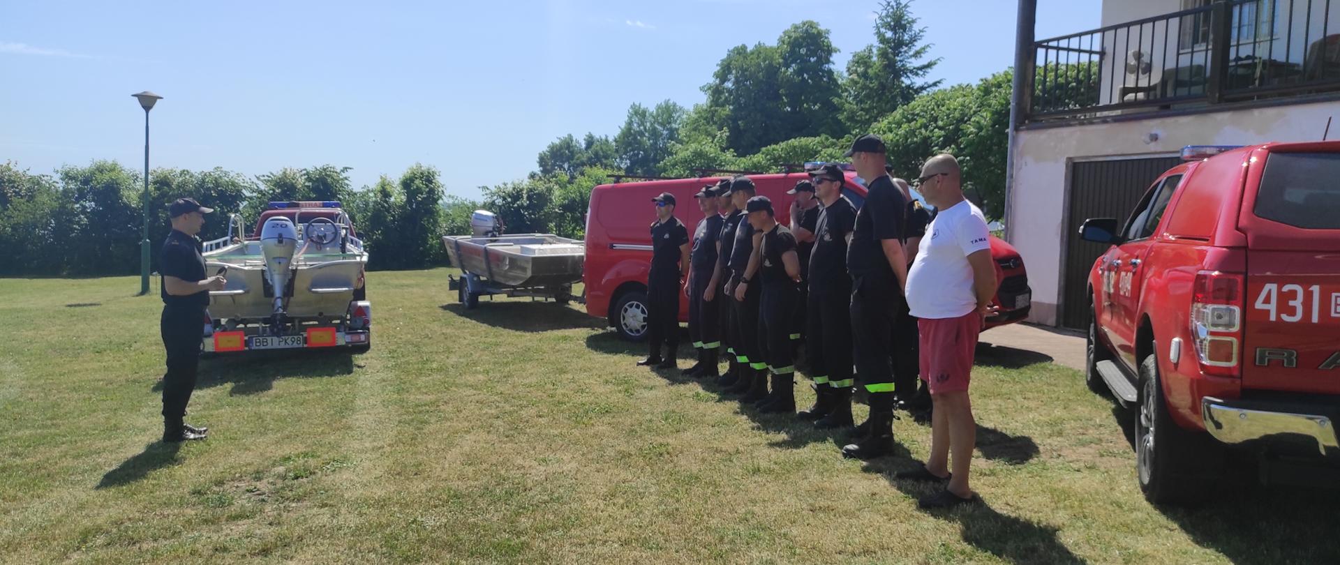 Na zielonej trawie przed czerwonymi wozami strażackimi do których podczepione są łódki stoją strażacy w dwuszeregu w czarnych mundurach.