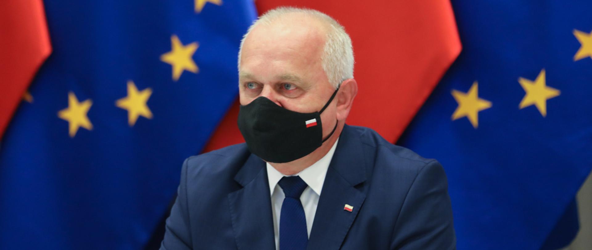 Wojewoda siedzi na tle flagi Polski oraz Unii Europejskiej. Na twarzy ma czarą maseczkę z biało-czerwoną flagą