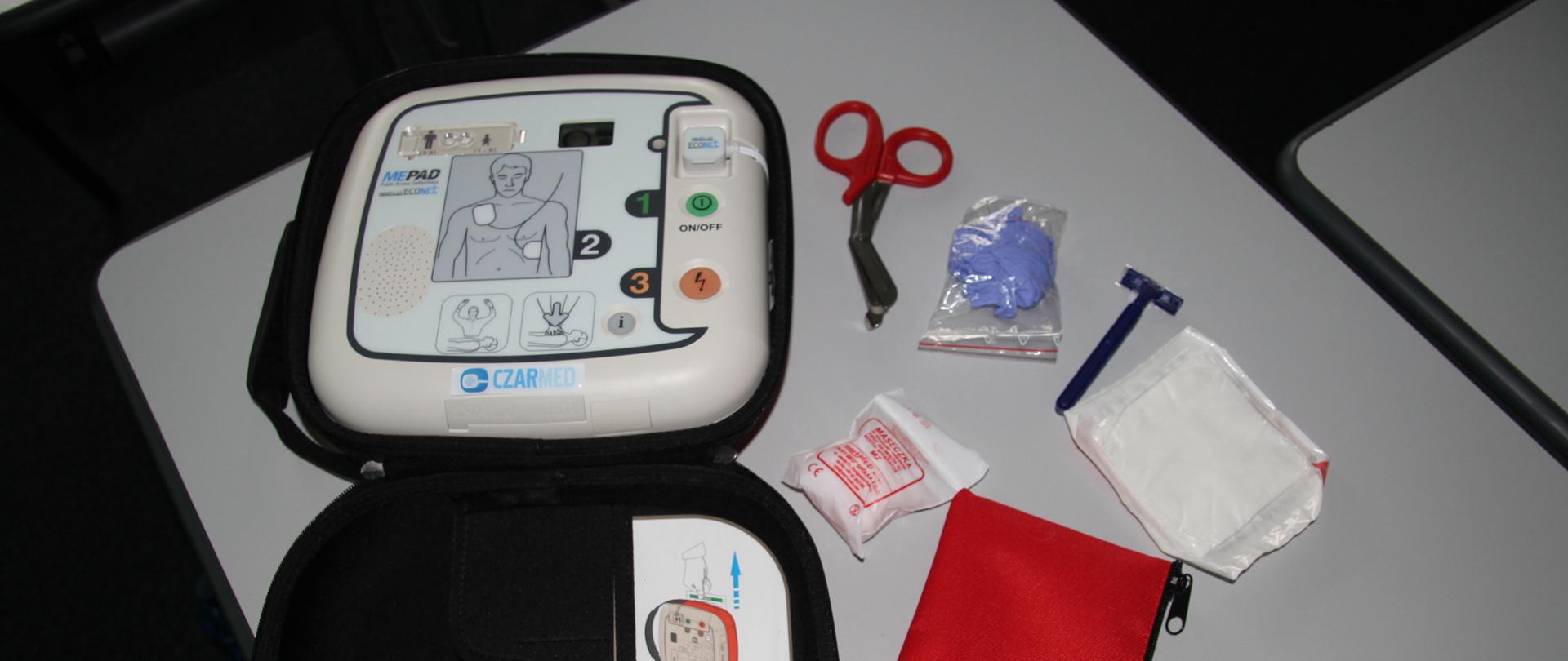 na stole widoczne jest białe urządzenie z rysunkiem człowieka, obok leży saszetka koloru czerwonego , gazik , nożyczki rękawiczki lateksowe oraz maszynka do golenia