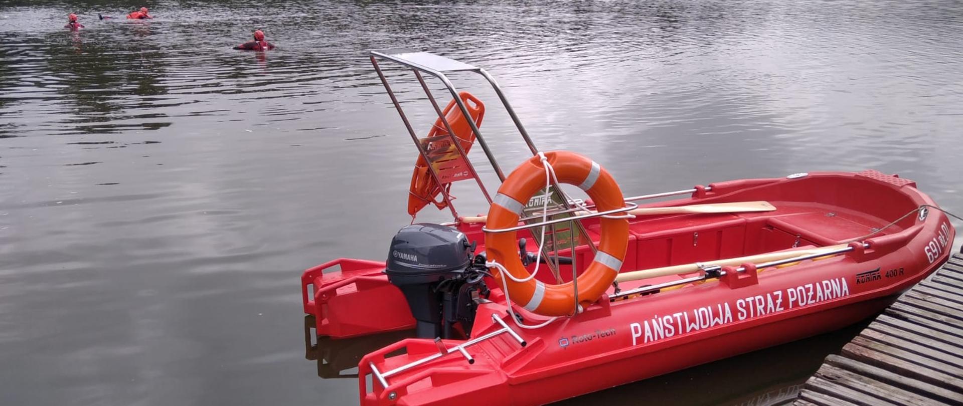 Łódka ratownicza przycumowana do pomostu. Na środku stawu trzech strażaków. w Tle las.