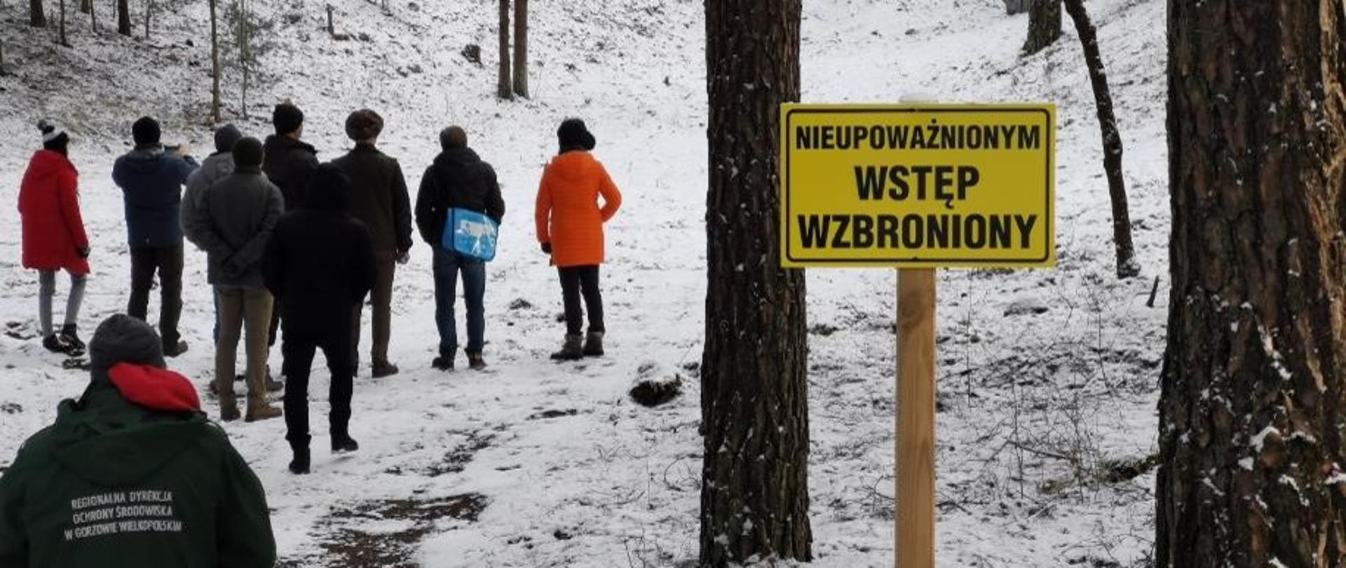 Zaśnieżony las, po lewej stronie na ścieżce stoi grupa kilkunastu osób. Na pierwszym planie po prawej stornie znajduje się żółta tablica z napisem: Nieupoważnionym wstęp wzbroniony.