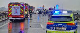 Zdjęcie przedstawia miejsce wypadku drogowego na mokrej, wielopasmowej trasie przelotowej. Na pierwszym planie po prawej stronie stoi policyjny radiowóz kombi w barwach srebrno-niebiesko-żółtych z włączonymi niebieskimi sygnałami błyskowymi. Po lewej stronie widoczne są dwa czerwone wozy strażackie z włączonymi światłami ostrzegawczymi, a na jezdni w tle stoi karetka pogotowia oraz rozbity biały samochód osobowy. Na drodze, wokół uszkodzonych pojazdów i rozrzuconych odłamków, pracują strażacy w ubraniach specjalnych i hełmach, policjanci oraz ratownicy medyczni. Niebo jest pochmurne, a mokra nawierzchnia odbija światła pojazdów ratowniczych.