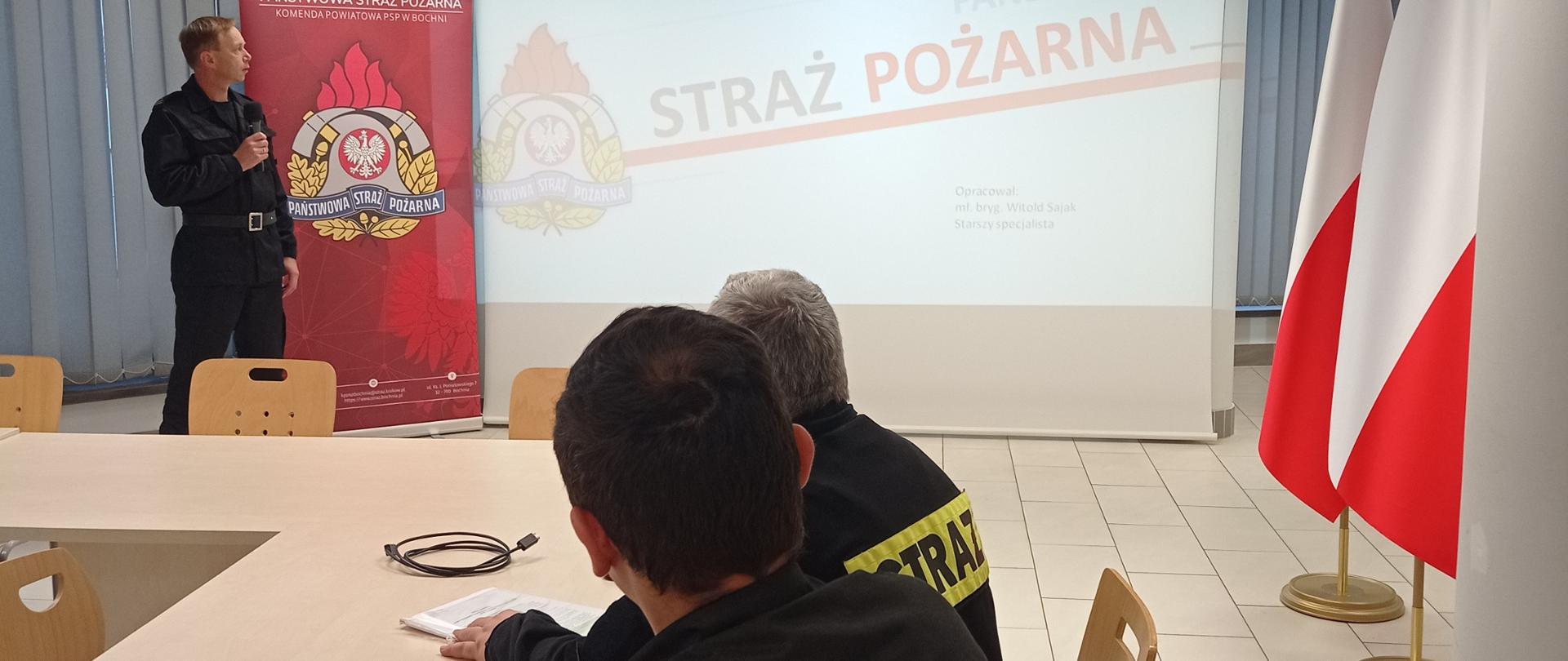 Specjalista BHP w służbowym mundurze strażackim prowadzi wykład z zakresu BHP na sali konferencyjnej. Słuchaczami są umundurowani naczelnicy jednostek OSP. W tle znajduje się ekran z prezentacją multimedialną i duże logo Państwowej straży Pożarnej