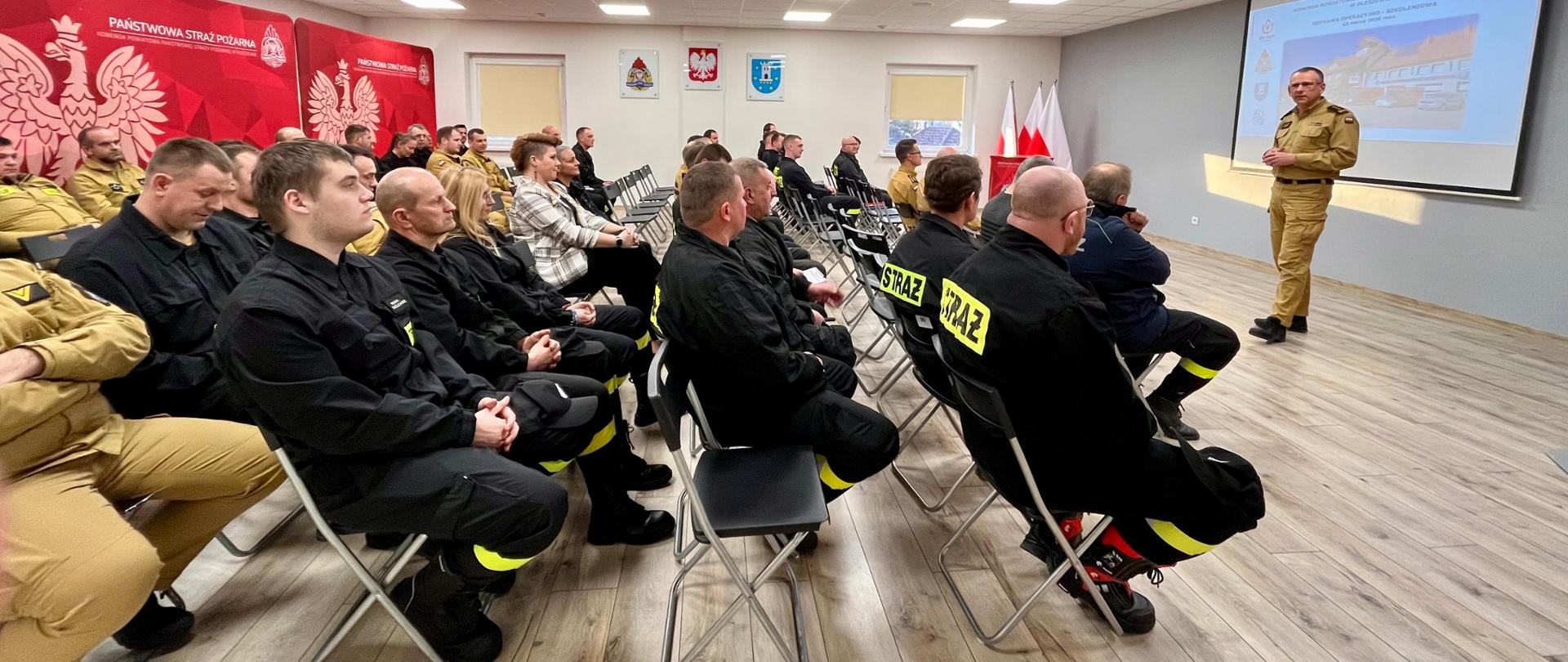 grupa strażaków w mundurach koszarowych siedzących na sali, w tle jedna osoba przemawia na stojąco