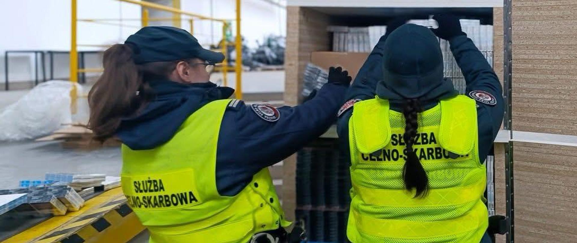 Dwie osoby w kamizelkach z napisem „Służba Celno‑Skarbowa” stoją przy metalowym regale i sprawdzają ułożone na półkach opakowania; w tle widać elementy hali magazynowej oraz żółtą barierkę.