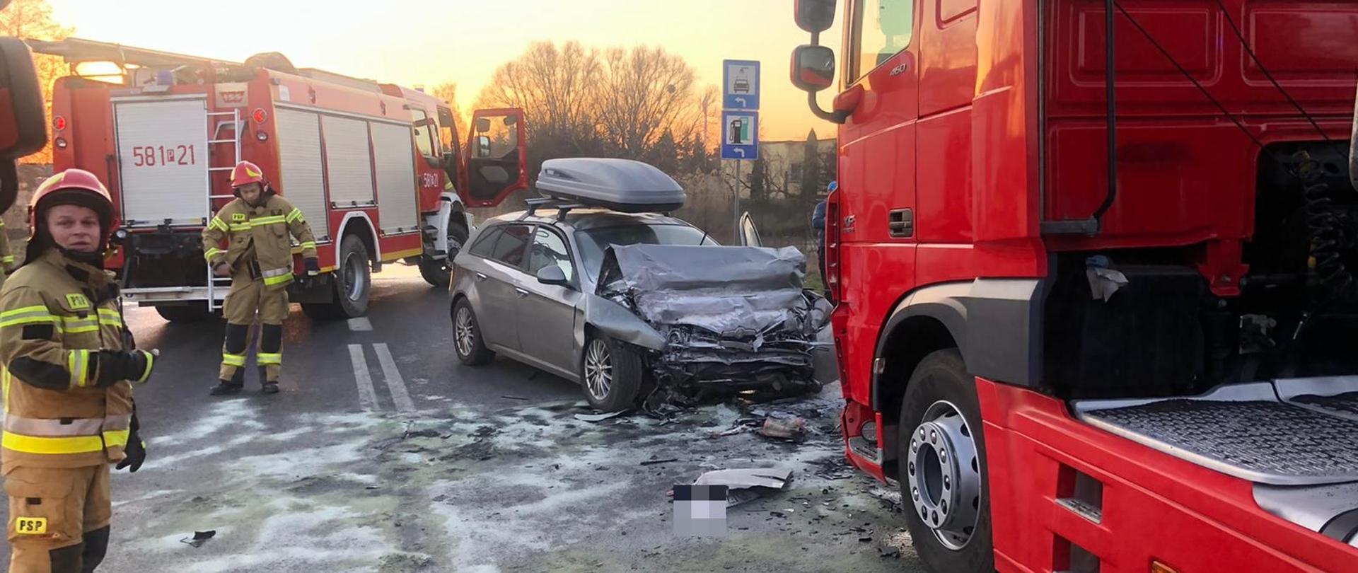 Czerwony samochód ciężarowy stoi z uszkodzonym przodem przed nim samochód osobowy srebrny z bagażnikiem na dachu z uszkodzonym przodem pomiędzy pojazdami plama posypana sorbentem wokół dwóch strażaków za nim czerwony samochód ciężarowy straży pożarnej 