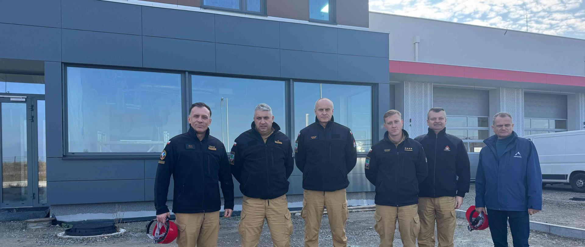 Do zdjęcia pozuje pięciu strażaków w piaskowych mundurach służbowych z czarnymi polarami oraz jeden mężczyzna w granatowej kurtce. Stoją na kamienistej ziemi przed nowym budynkiem.