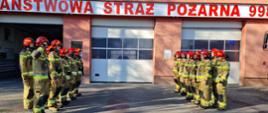 Strażacy stoją naprzeciw siebie w w dwuszeregu po każdej stronie na placu przed garażami. Dowódcy salutują, a reszta strażaków stoi w pozycji zasadniczej.