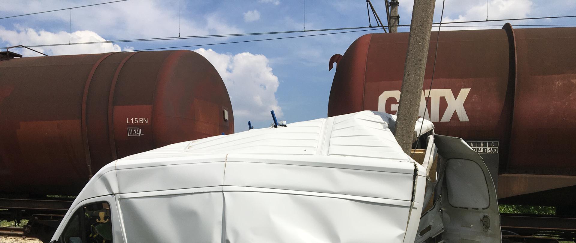 Słonecznie. Biały samochód dostawczy z uszkodzoną kabiną, bokiem oraz tyłem po zderzeniu z pociągiem towarowym stoi na nasypie kolejowym, porośniętym zielonym trawą. Obok brązowe cysterny kolejowe pociągu towarowego.