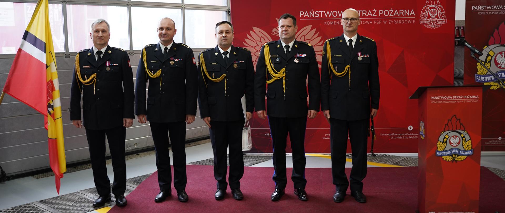 Do zdjęcia pozują Mazowiecki Komendant Wojewódzki PSP nadbryg. Artur Gonera wraz z dwoma zastępcami oraz zdający i przyjmujący obowiązki Komendanta Powiatowego PSP w Żyrardowie. Są w czarnych mundurach wyjściowych. Stoją na bordowym dywanie przed czerwoną ścianką z napisem „PAŃSTWOWA STRAŻ POŻARNA | KOMENDA POWIATOWA PSP W ŻYRARDOWIE”. Zdjęcie zrobione na hali garażowej.