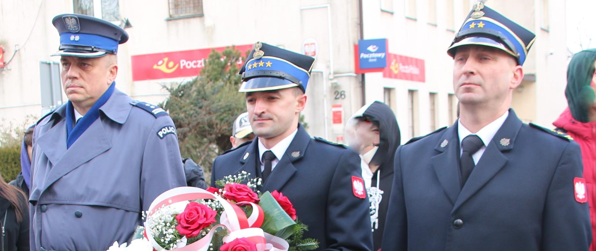 Na pierwszym palnie widnieje postać Komendanta Powiatowego Państwowej Straży Pożarnej w Jędrzejowie w umundurowaniu galowym oraz płaszczu. Na głowie ma założoną czapkę rogatywkę. W rękach trzyma wiązankę biało- czerwonych kwiatów. Po jego lewej stronie znajduje się zastępca komendanta PSP także w umundurowaniu galowym oraz płaszczu, a po jego prawej- Komendant Powiatowy Policji.