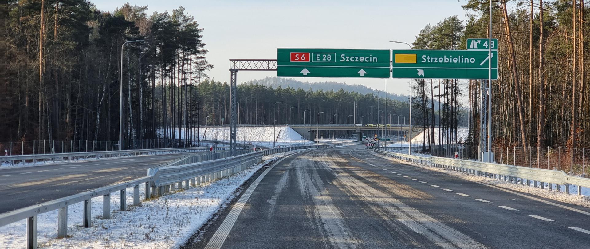 Zdjęcie przedstawia drogę ekspresową S6 w pobliżu węzła Strzebielino. Na pierwszym planie jezdnia prowadząca do Szczecina. Nad drogą wisi tablica drogowskazowa na Szczecin i Strzebielino, a na drugim planie widać wiadukt na węźle.
