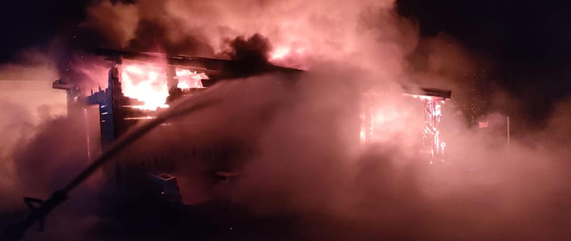 Na zdjęciu widać budynek drewniany całkowicie objęty pożarem. Jest duże zadymienie. Na budynek z węza strażackiego lana jest woda. Jest ciemno.