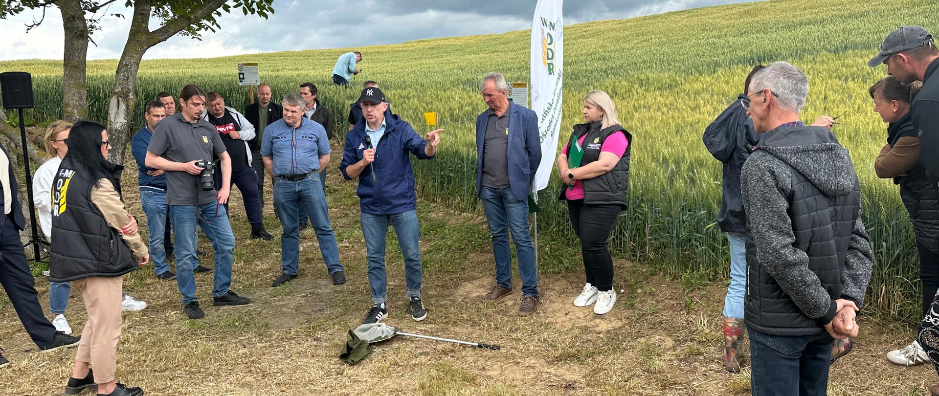  Mężczyzna stoi z mikrofonem na leżącym przed nim czerpakiem entomologicznym. Dookoła stoi grupa słuchających go osób, w tle plantacja zbóż