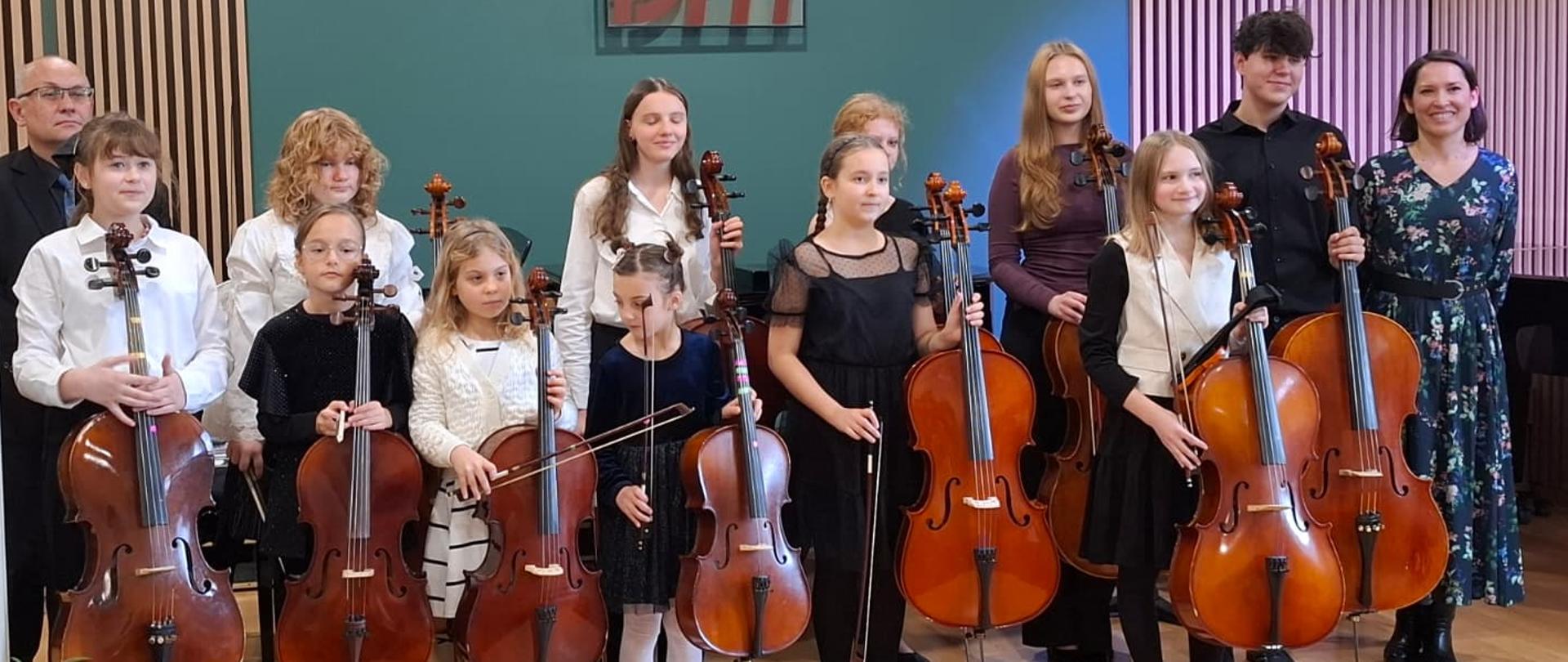 Uczniowie z wiolonczelami w rękach pozują do zdjęcia na scenie w sali koncertowej, po lewej stronie stoi akompaniator ubrany w czarną koszulę a po prawej stronie stoi pani od wiolonczeli która zapowiadała koncert ubrana w ciemną sukienkę w kwiatki. Uczniowie są wszyscy w galowych strojach. W tle widoczna jest Zielona ściana z drewnianymi lamelami.