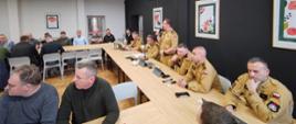 Spotkanie w sali konferencyjnej: strażacy w mundurach siedzą przy długim stole, a jeden z nich przemawia do mikrofonu. Uczestnicy słuchają i prowadzą rozmowy, na stołach znajdują się laptopy, dokumenty oraz napoje. W tle widać innych uczestników oraz ściany z oprawionymi grafikami.