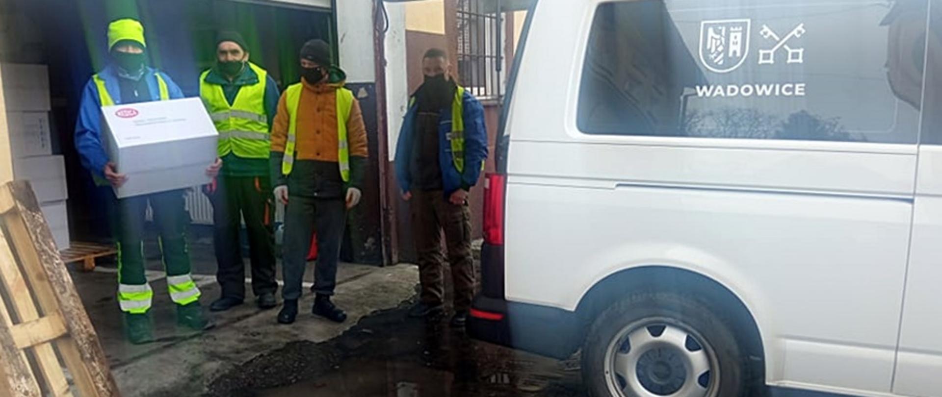 Czterej pracownicy samorządu lokalnego stoją przy samochodzie dostawczym, jeden z nich trzyma w rękach paczkę z maseczkami ochronnymi. 