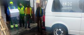 Czterej pracownicy samorządu lokalnego stoją przy samochodzie dostawczym, jeden z nich trzyma w rękach paczkę z maseczkami ochronnymi. 