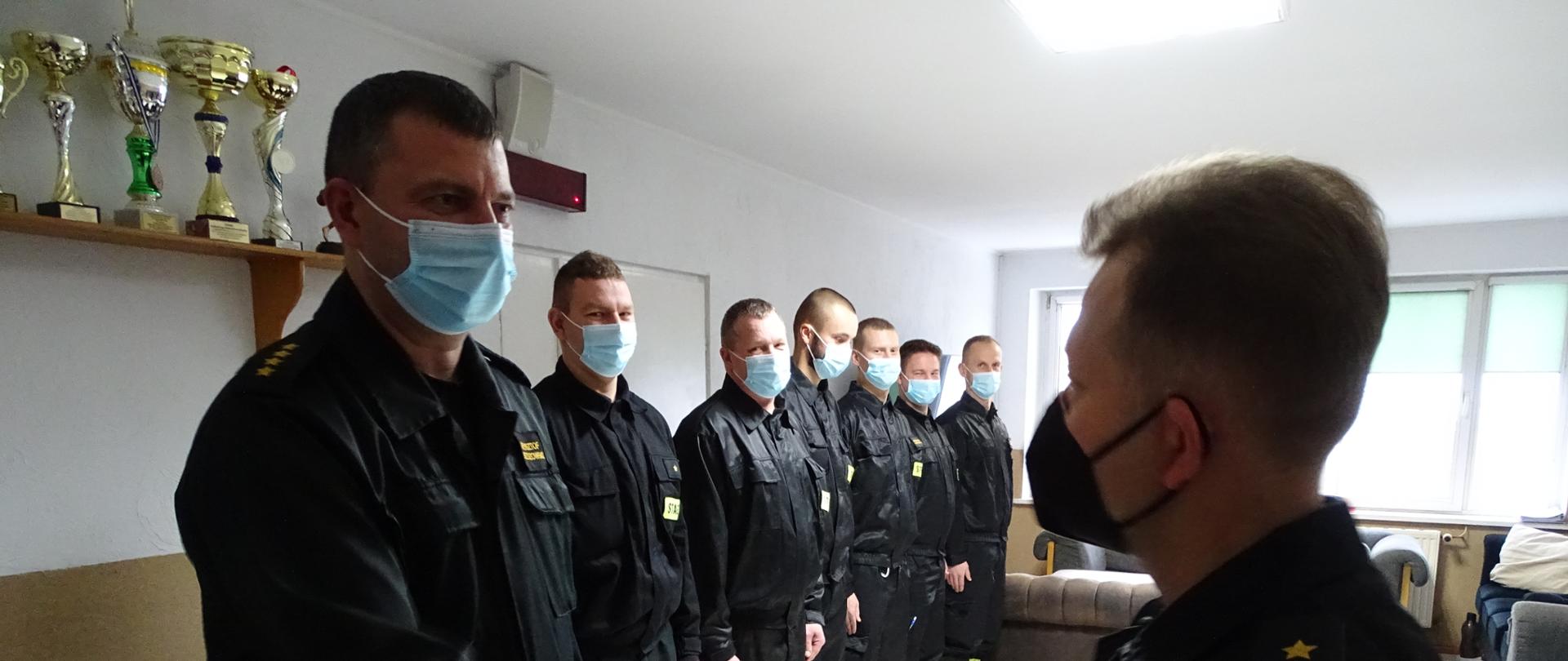 Zdjęcie wykonano w budynku Komendy Powiatowej Państwowej Straży Pożarnej w Sępólnie Krajeńskim w pomieszczeniu świetlicy, na zdjęciu starszy kapitan Krzysztof Brzozowski (po lewej) razem ze strażakami ze zmiany służbowej ubrani w mundury koszarowe i maseczki jednorazowe. Komendant Powiatowy Państwowej Straży Pożarnej starszy brygadier Maciej Suchomski (po prawej) gratuluje starszemu kapitanowi Krzysztofowi Brzozowskiemu. 