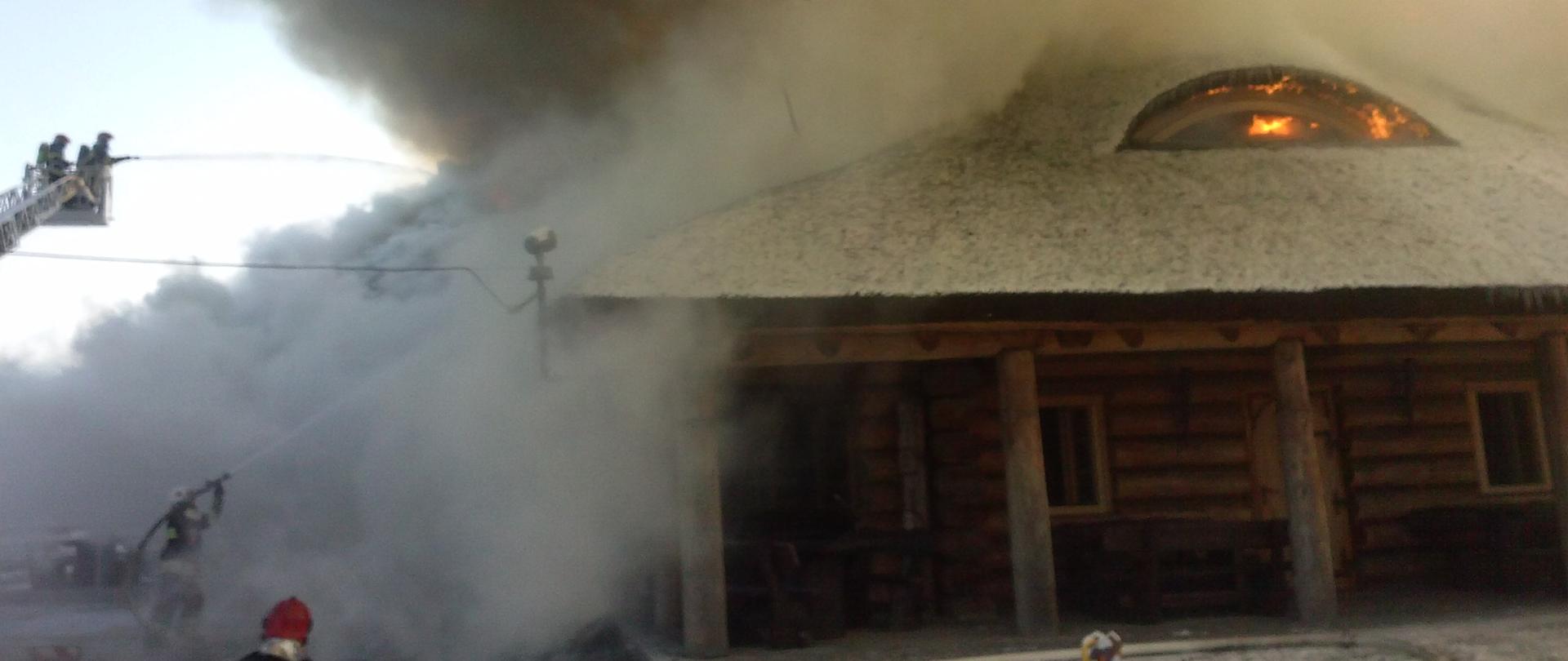 Po lewej stronie strażak gaszący palący się drewniany dom, kryty strzechą