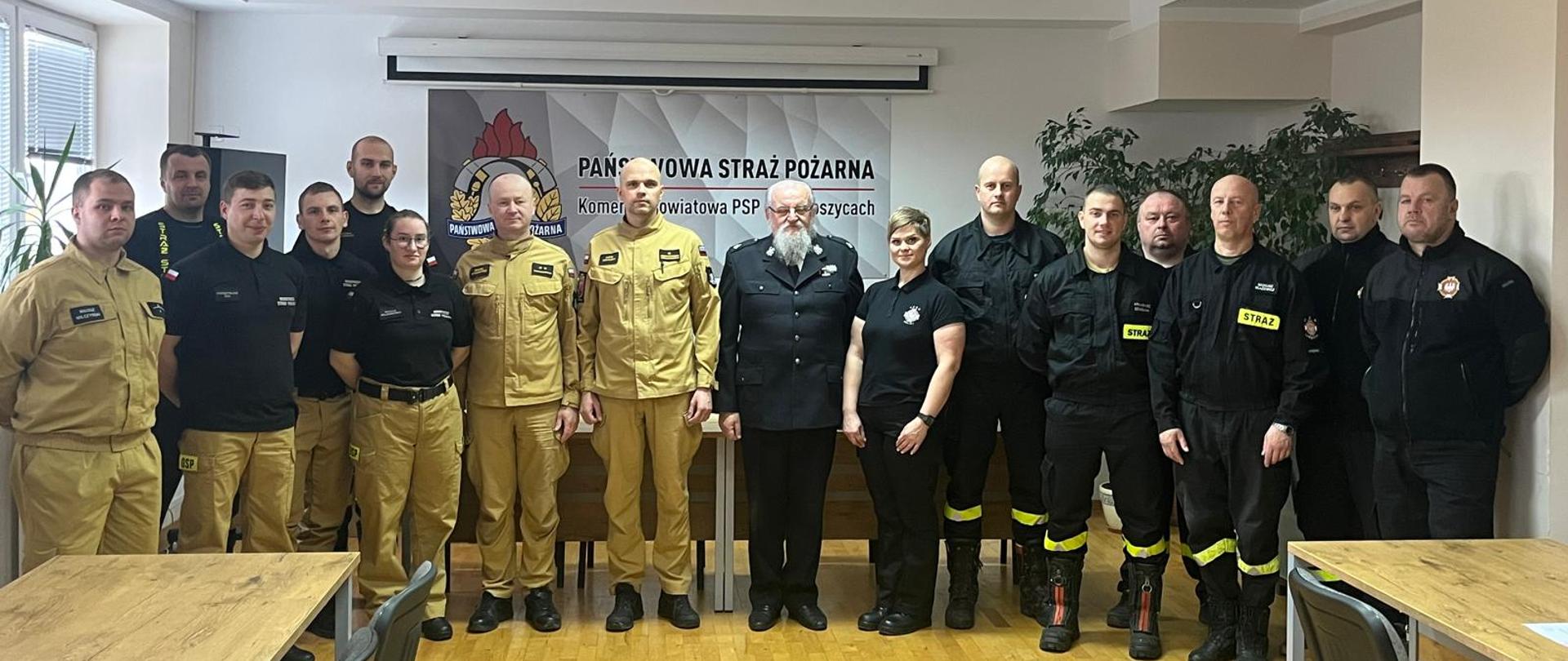 Grupa strażaków Państwowej Straży Pożarnej i Ochotniczej Straży Pożarnej stoi wspólnie w sali szkoleniowej. Za nimi znajduje się baner z napisem „Państwowa Straż Pożarna – Komenda Powiatowa PSP w Bartoszycach”. Uczestnicy ubrani są w umundurowanie służbowe w kolorze czarnym i piaskowym. Zdjęcie wykonane w pomieszczeniu z drewnianą podłogą i stołami ustawionymi po bokach.