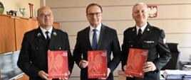 Trzej mężczyźni w garniturze i munudurach trzymają czerwone teczki przed sobą. Za nimi w tle biurko z krzesłem obrotowym i dokumentami na nim. Na ścianie godło.
