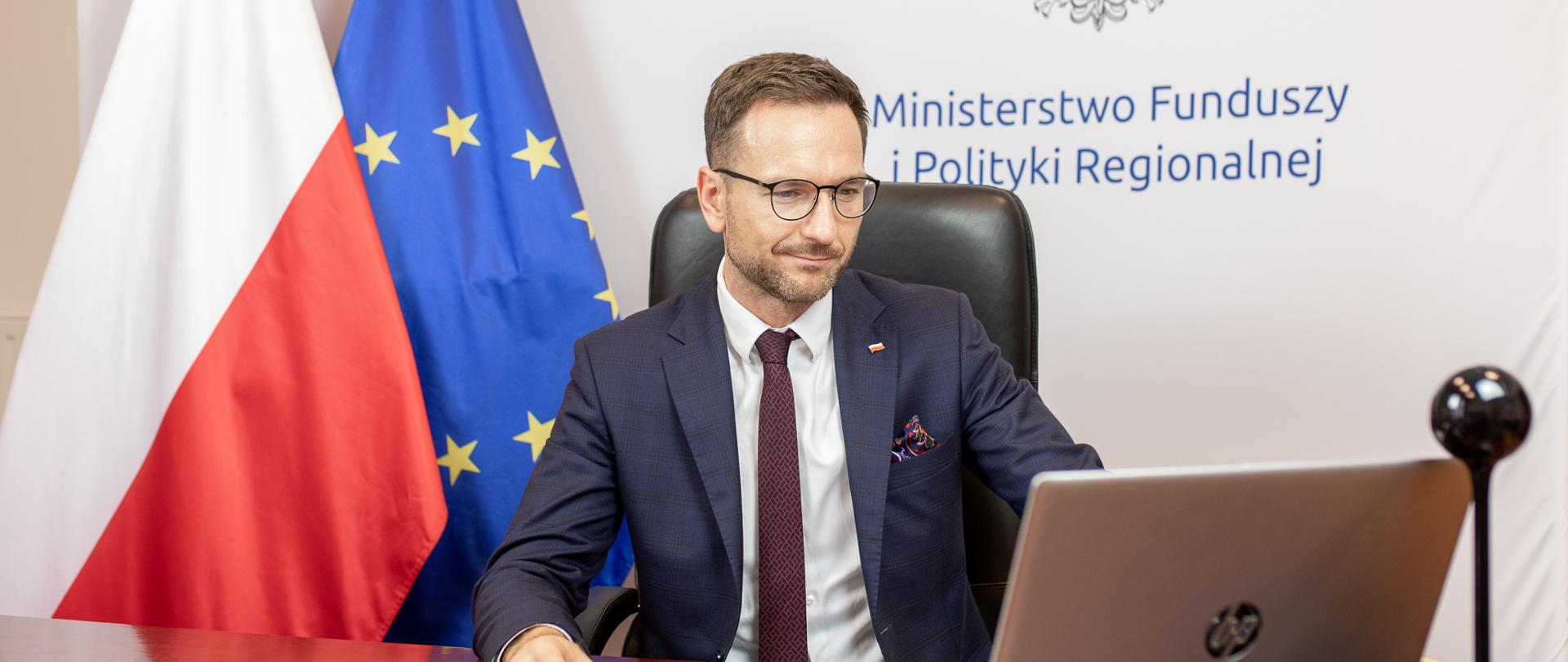 Minister Waldemar Buda przy biurku, w tle flaga Polski i Unii Europejskiej