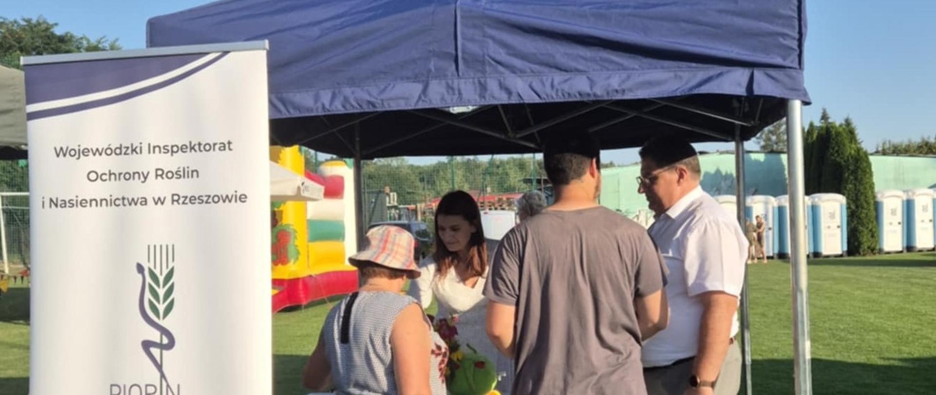 Trzy osoby stojące pod granatowym namiotem. Przed nimi stoi stół z białym obrusem. Po lewej stronie jest baner z logiem instytucji. 