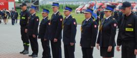 Na zdjęciu widoczni są strażacy podczas apelu z okazji dnia strażaka. Osoby stoją w szyku pojedynczym na placu wyłożonym kostką brukową. Z pośród strażaków widoczna jest jedna kobieta 2 osoby umundurowane w mundury koszarowe pozostali w mundury galowe.
