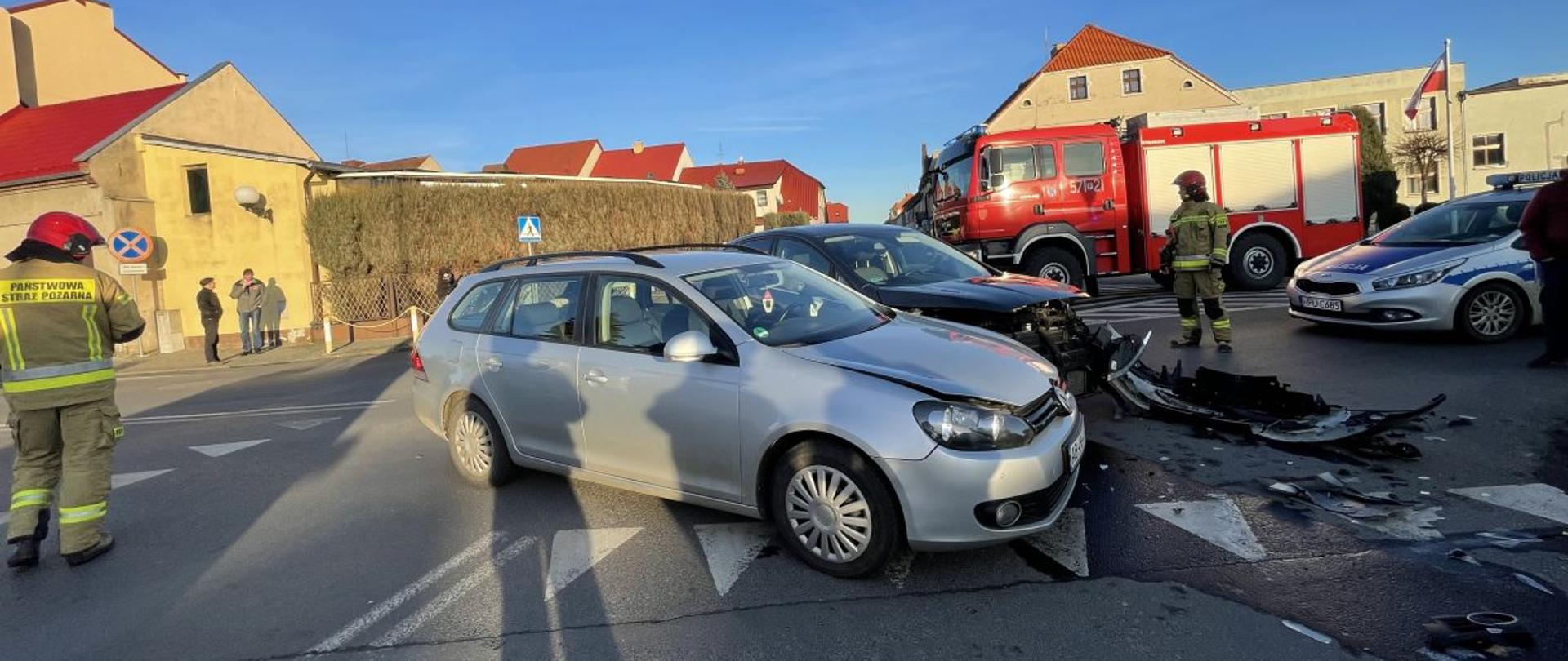 Kolizja w Miejskiej Górce. Droga krajowa nr 36. Na środku skrzyżowania stoją dwa uszkodzone samochody. Jeden jest koloru srebrnego, drugi ciemny. Na jezdni widoczna plama płynów eksploatacyjnych oraz oderwane elementy pojazdów. Wokół aut stoi dwóch strażaków w czerwonych hełmach. W tle wóz strażacki, radiowóz policyjny, maszt z flagą i budynki.