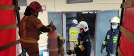 Na pierwszym planie strażak przemieszczający butle z tlenem na pojeździe pożarniczym, na drugim palnie tlenownia i rampa rozładunkowa